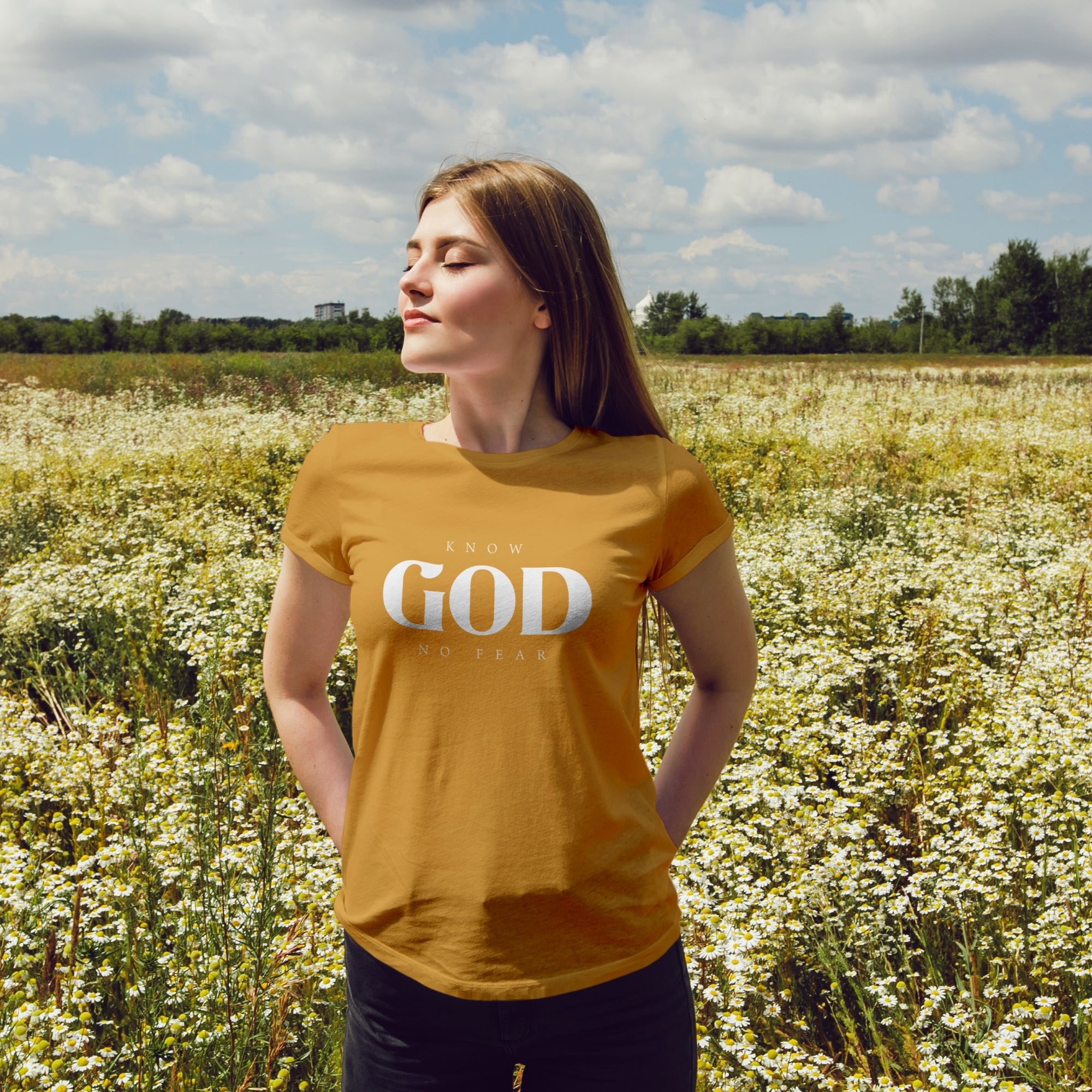 A woman standing confidently in a field of wildflowers, wearing a mustard-colored t-shirt with the text 'Know God, No Fear,' radiating faith and serenity.